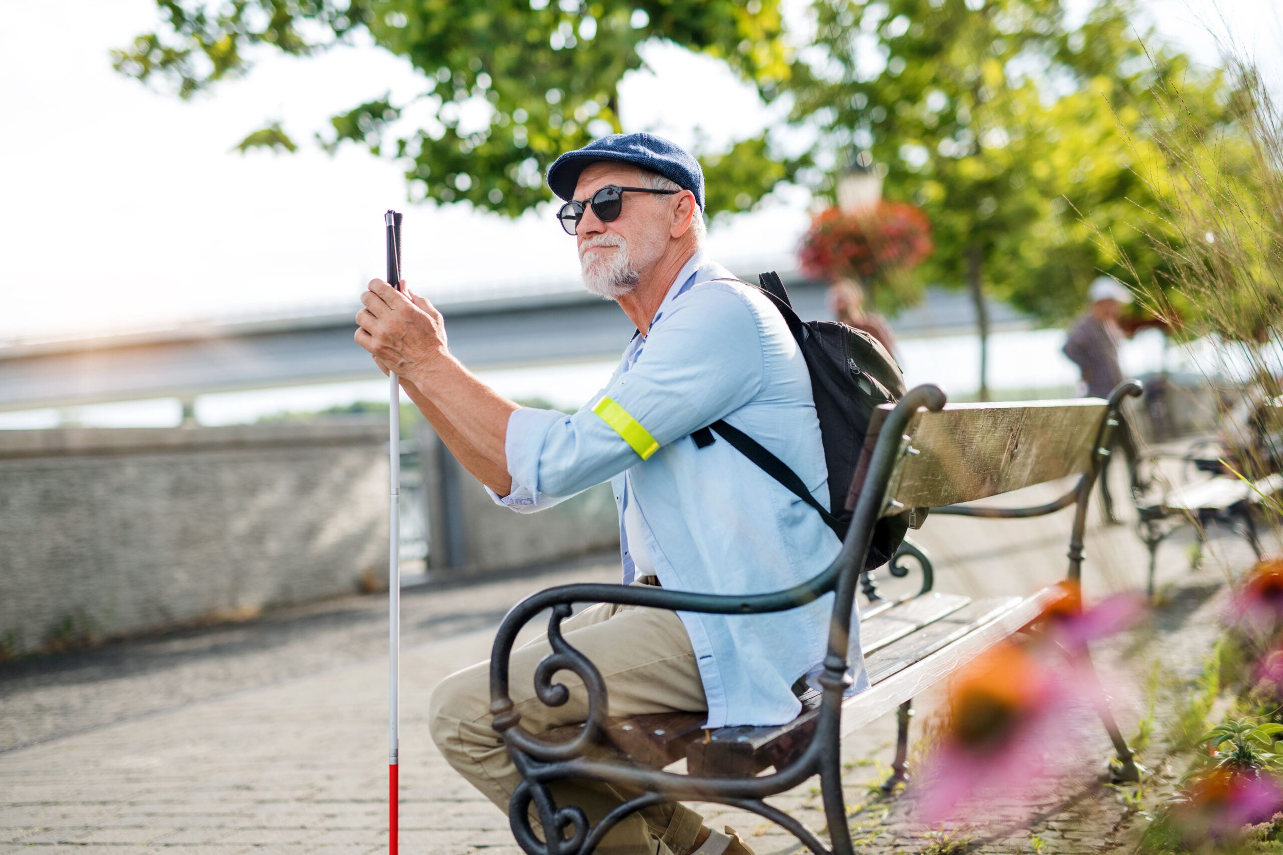 Senior blind man with cane sitting on bench in park in city.