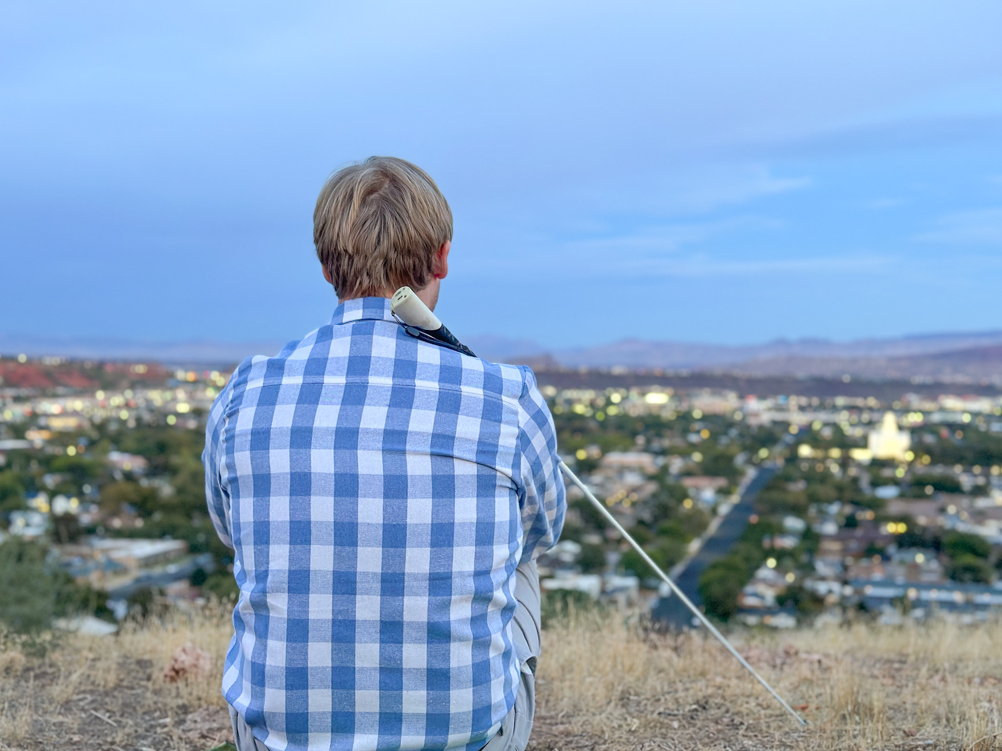 Man wearing a blue plaid shirt sits on a hillside overlooking a city at dusk, holding a white cane beside him as city lights glow in the distance.